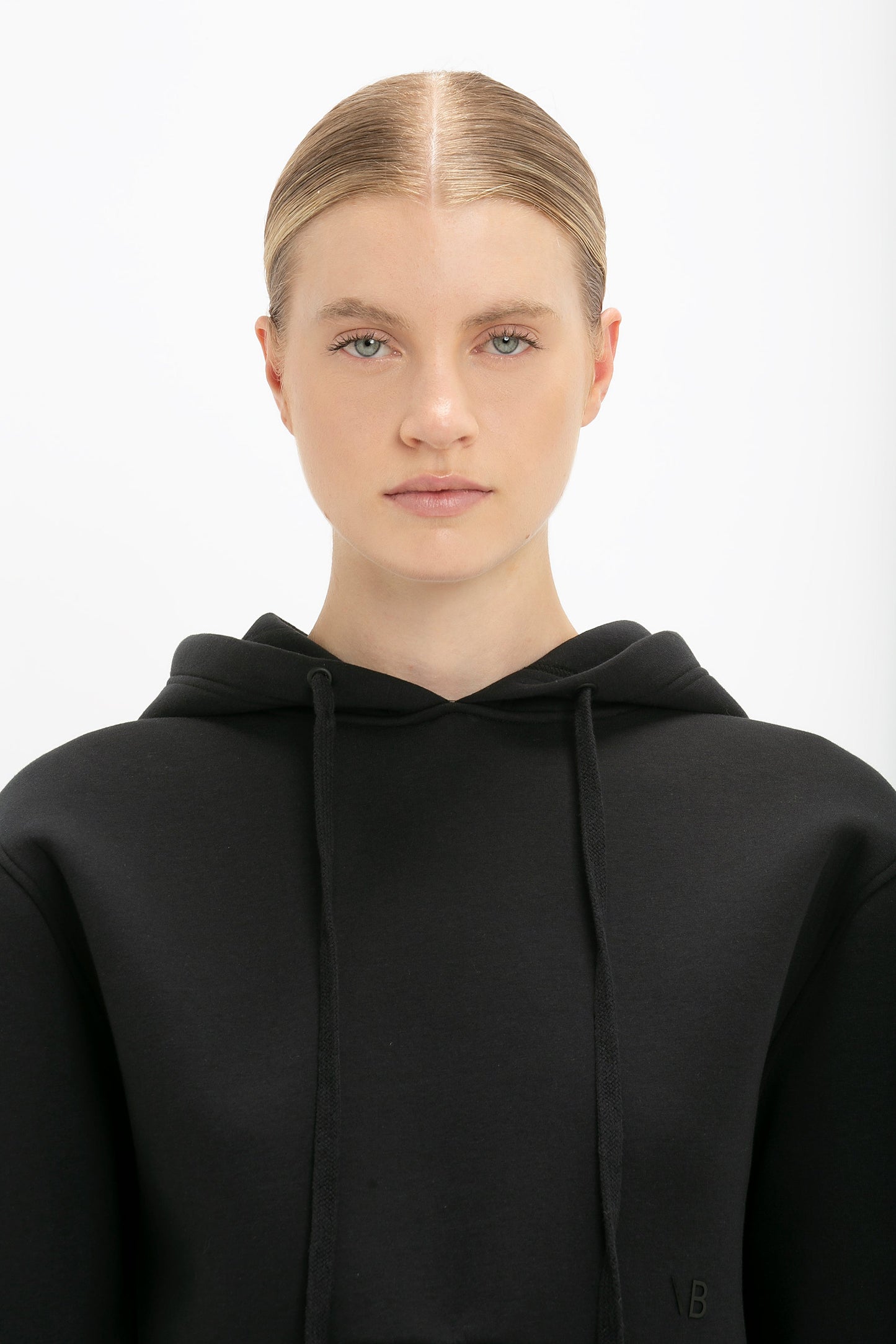 A person with a neutral expression wearing a Cropped Neoprene Hoodie In Black, reminiscent of Victoria Beckham's minimalist style, stands against a white background.