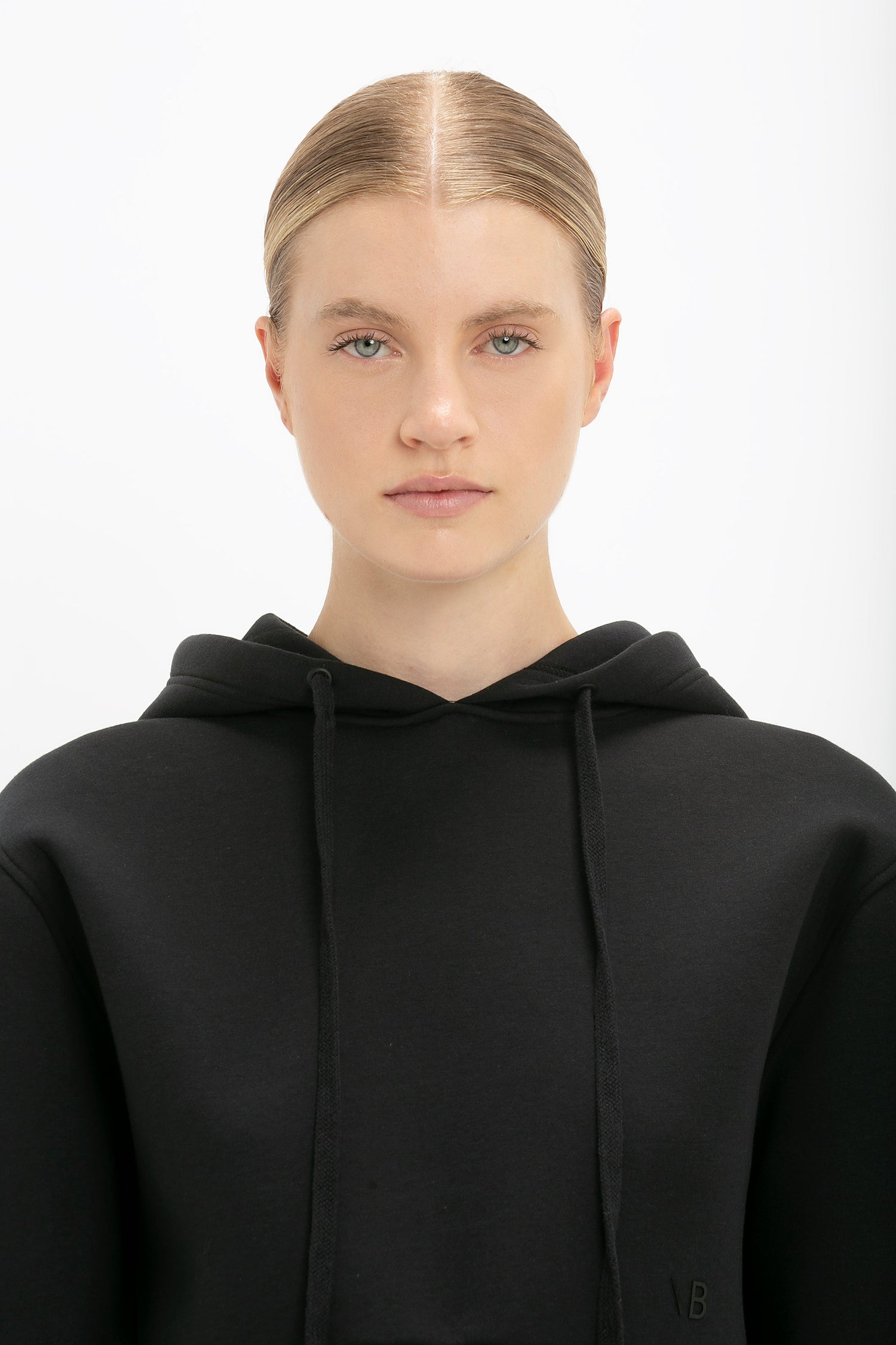 A person with a neutral expression wearing a Cropped Neoprene Hoodie In Black, reminiscent of Victoria Beckham's minimalist style, stands against a white background.