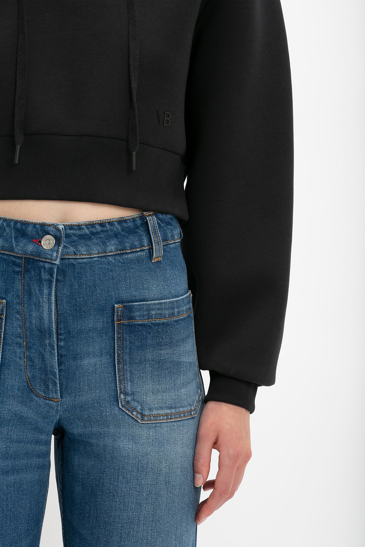 A person wearing a Victoria Beckham Cropped Neoprene Hoodie In Black and blue jeans with front pockets is shown from the shoulders to the waist.