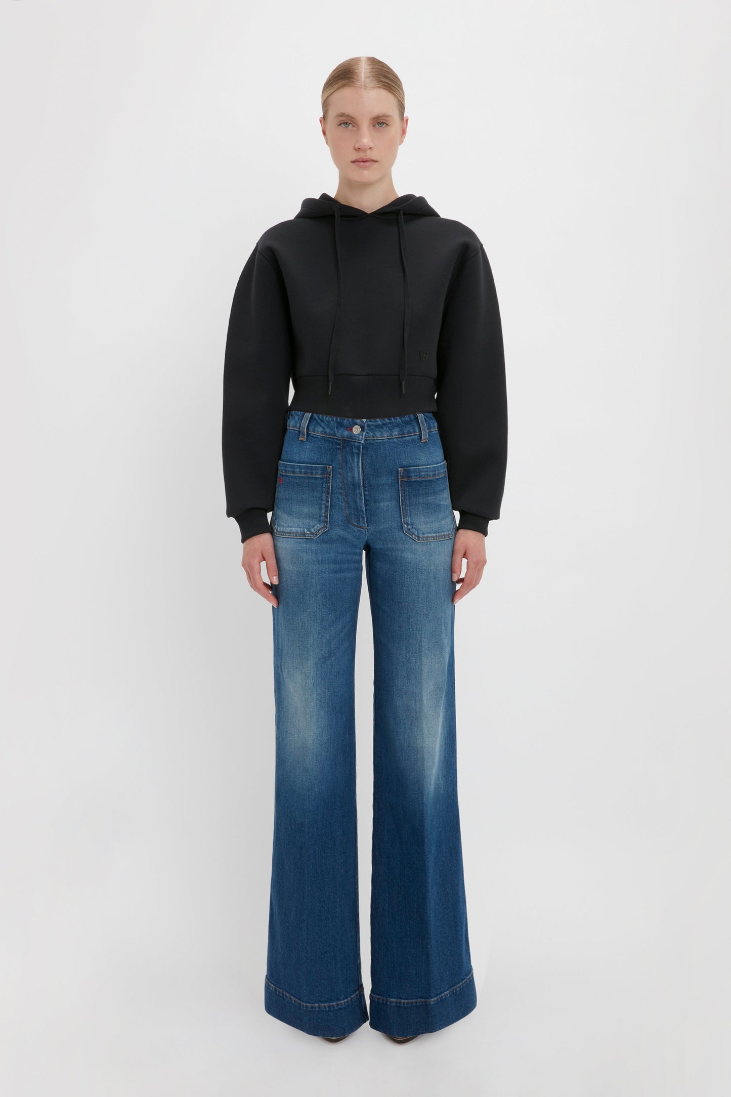 A person stands against a plain background, wearing a stylish Cropped Neoprene Hoodie In Black by Victoria Beckham and wide-leg blue jeans with front pockets.
