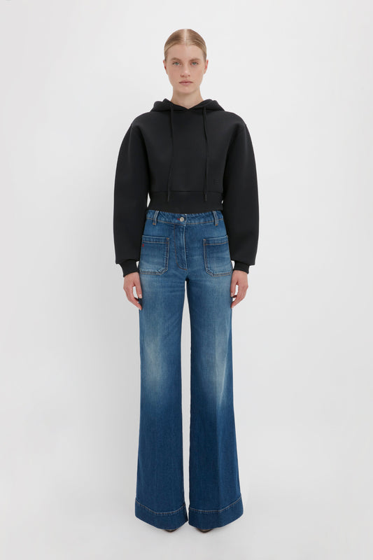 A person stands against a plain background, wearing a stylish Cropped Neoprene Hoodie In Black by Victoria Beckham and wide-leg blue jeans with front pockets.