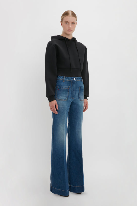 A person stands against a plain background wearing a black Victoria Beckham Cropped Neoprene Hoodie In Black and blue high-waisted, wide-leg Alina Jeans.