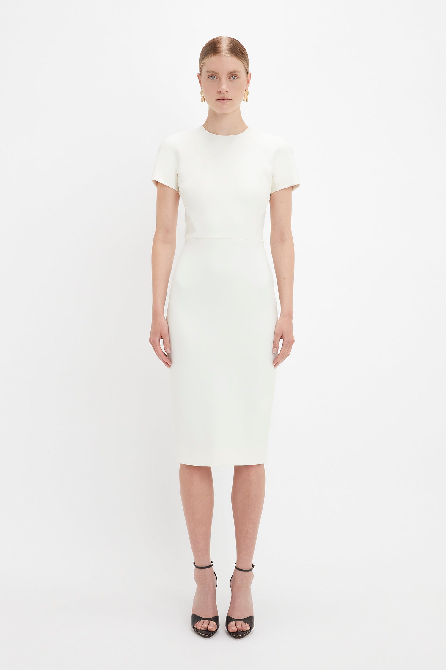 A woman in a Victoria Beckham fitted T-shirt dress in ivory and black high-heeled sandals stands against a stark white background.