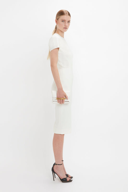 A woman in a sleek white Victoria Beckham fitted T-shirt dress with short sleeves holds a mini chain pouch and wears black pointy toe stilettos, standing against a plain white background.