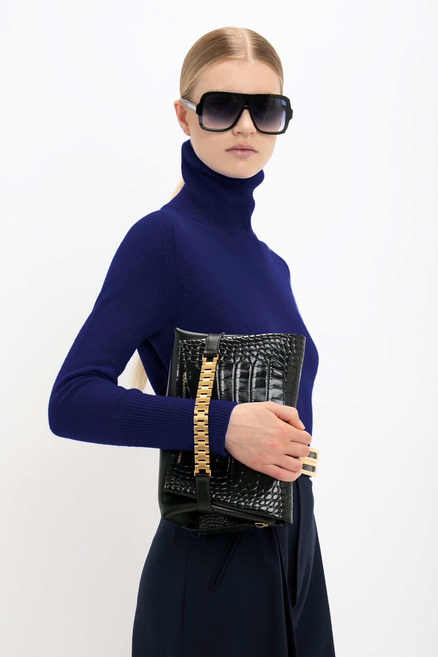 A person wearing a Polo Neck Jumper In Navy by Victoria Beckham with large sunglasses holds a black textured handbag with a gold chain detail.