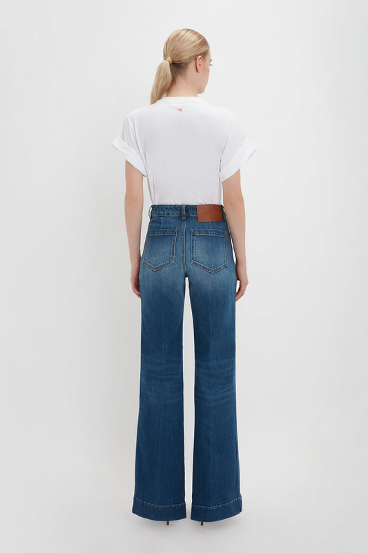 Woman in Victoria Beckham's white asymmetric relaxed fit T-shirt and blue flared jeans with a brown belt, standing against a white background, viewed from behind.