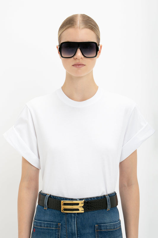 Woman in oversized Victoria Beckham white T-shirt and jeans with a black belt, wearing oversized black sunglasses, standing against a white background.