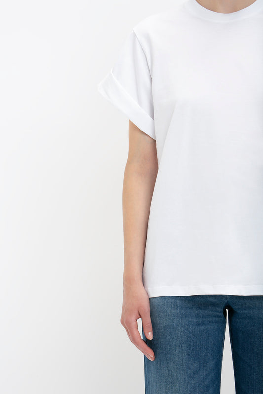 A person wearing an oversized white Asymmetric Relaxed Fit T-shirt with the Victoria Beckham monogram and blue jeans, cropped at the neck, focusing on the torso and arms.