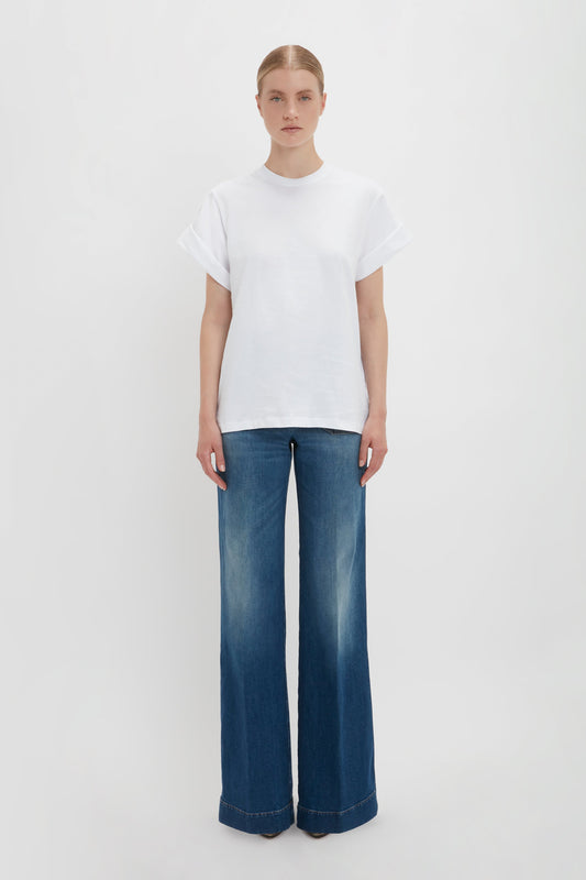 A woman stands against a white background wearing an oversized Victoria Beckham Asymmetric Relaxed Fit T-Shirt in White and blue Alina jeans.