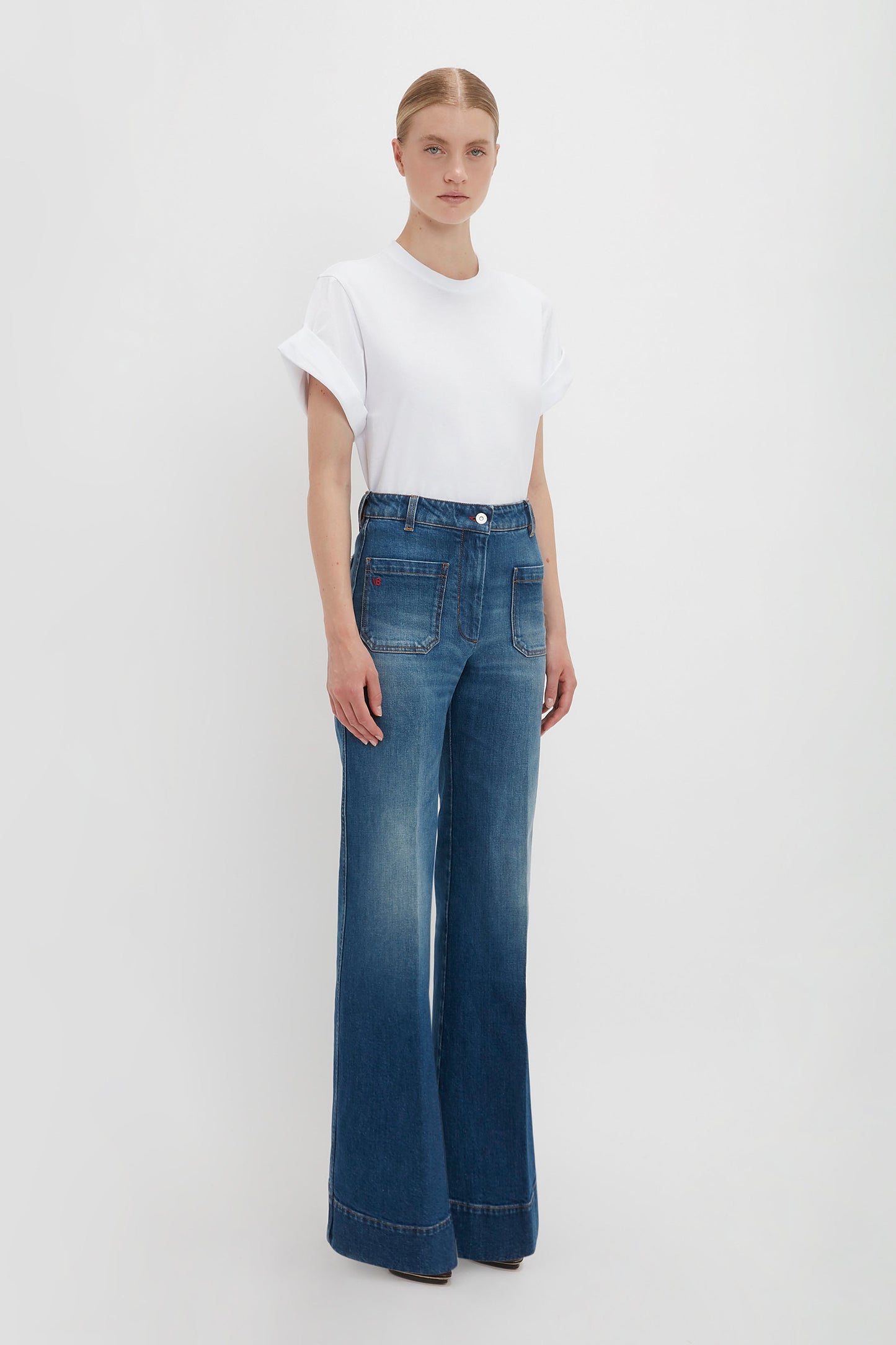 Woman in a Victoria Beckham Asymmetric Relaxed Fit T-Shirt in White and blue high-waisted jeans standing against a white background.