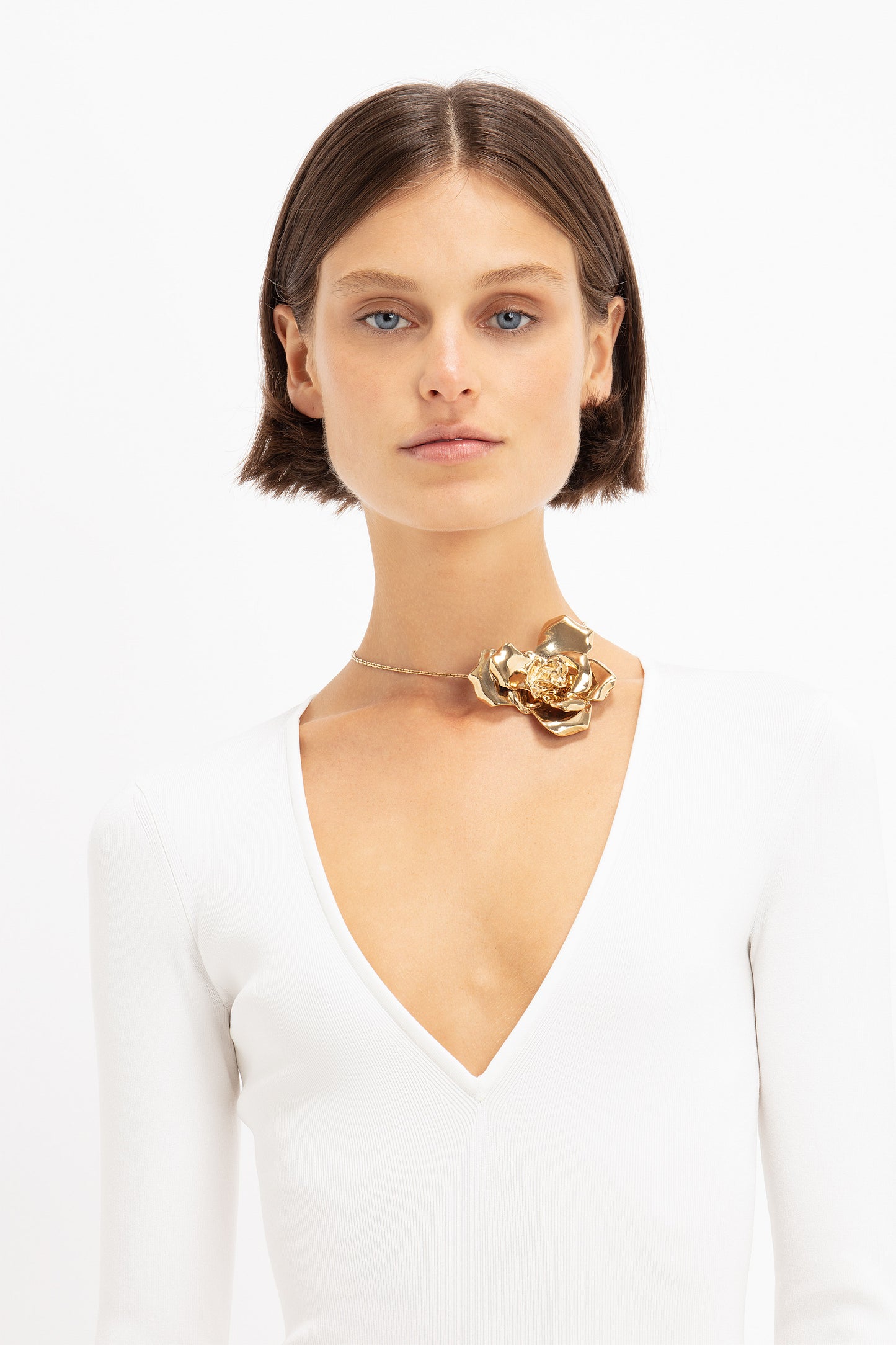 A person with short brown hair wears a white V-neck top and a statement accessory: the Exclusive Flower Necklace In Gold by Victoria Beckham, against a plain white background.