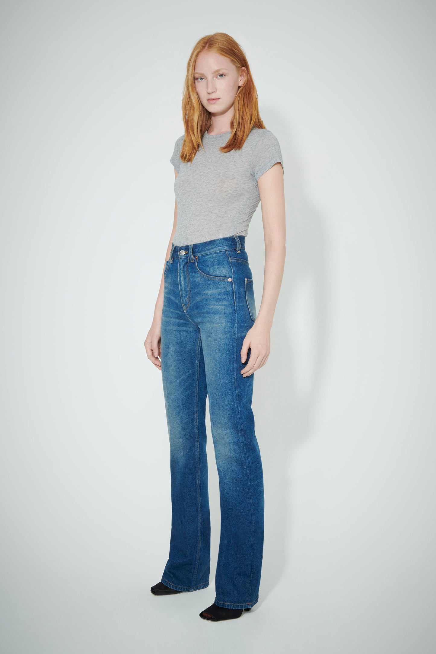 A woman with long, red hair wearing a T-Shirt Bodysuit in Grey by Victoria Beckham and blue jeans stands against a plain background. Her outfit is complemented by the bodysuit, showcasing her sense of style.