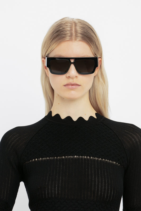 A woman with blonde hair wearing Victoria Beckham V Plaque Frame Sunglasses In Black with a double bridge and a textured black top, standing against a white background.