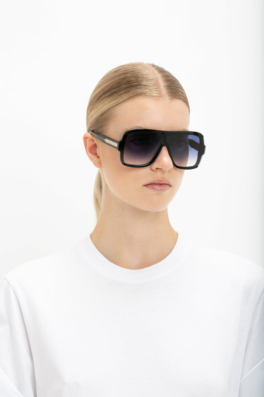 A woman with blonde hair slicked back, wearing Victoria Beckham Layered Mask Sunglasses In Black Gradient, and a plain white t-shirt, against a white background.