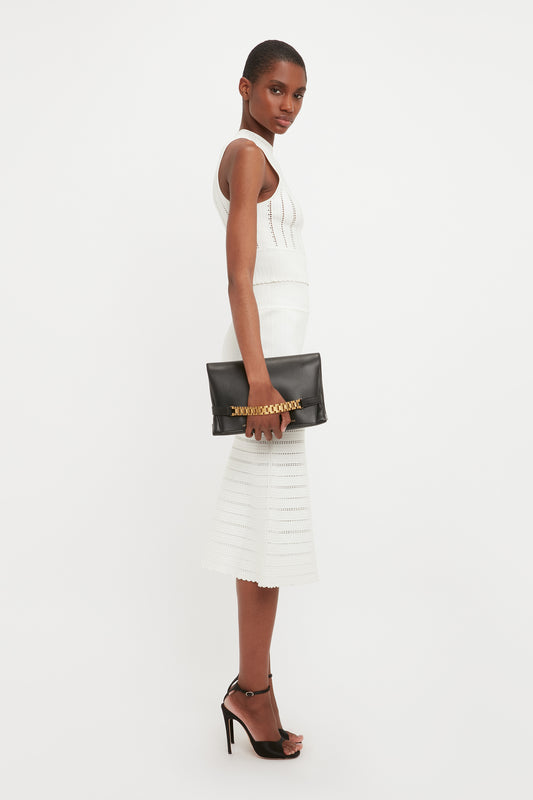 A woman in a white sleeveless knit dress with a flared midi skirt holds a black Victoria Beckham chain pouch bag and stands in a white studio. She looks over her shoulder with subtle makeup and short hair.