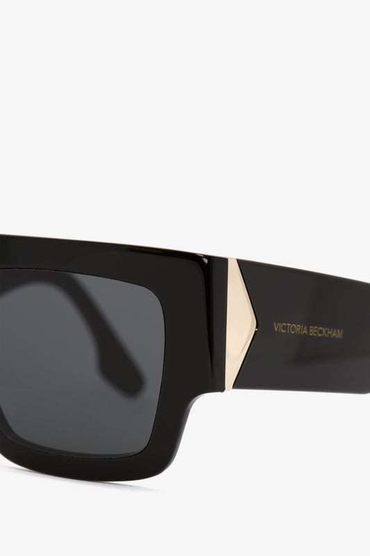 Close-up of black rectangular Victoria Beckham V Plaque Frame Sunglasses with a gold accent on the temple, isolated on a white background.