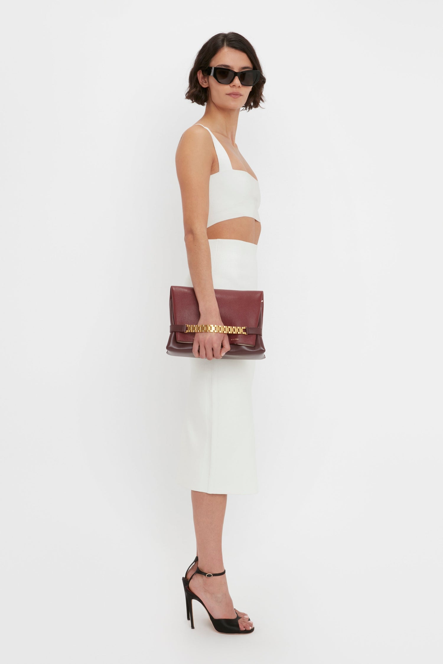 A woman in a Victoria Beckham VB Body Strap Bandeau Top In White and VB Body Fitted Midi Skirt, holding a maroon handbag, wearing sunglasses and black high heels, posing against a white background.