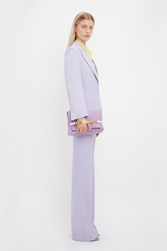 A woman in a lavender suit and yellow blouse stands sideways, holding a Victoria Beckham Chain Pouch with Strap in Lilac Suede, against a white background.