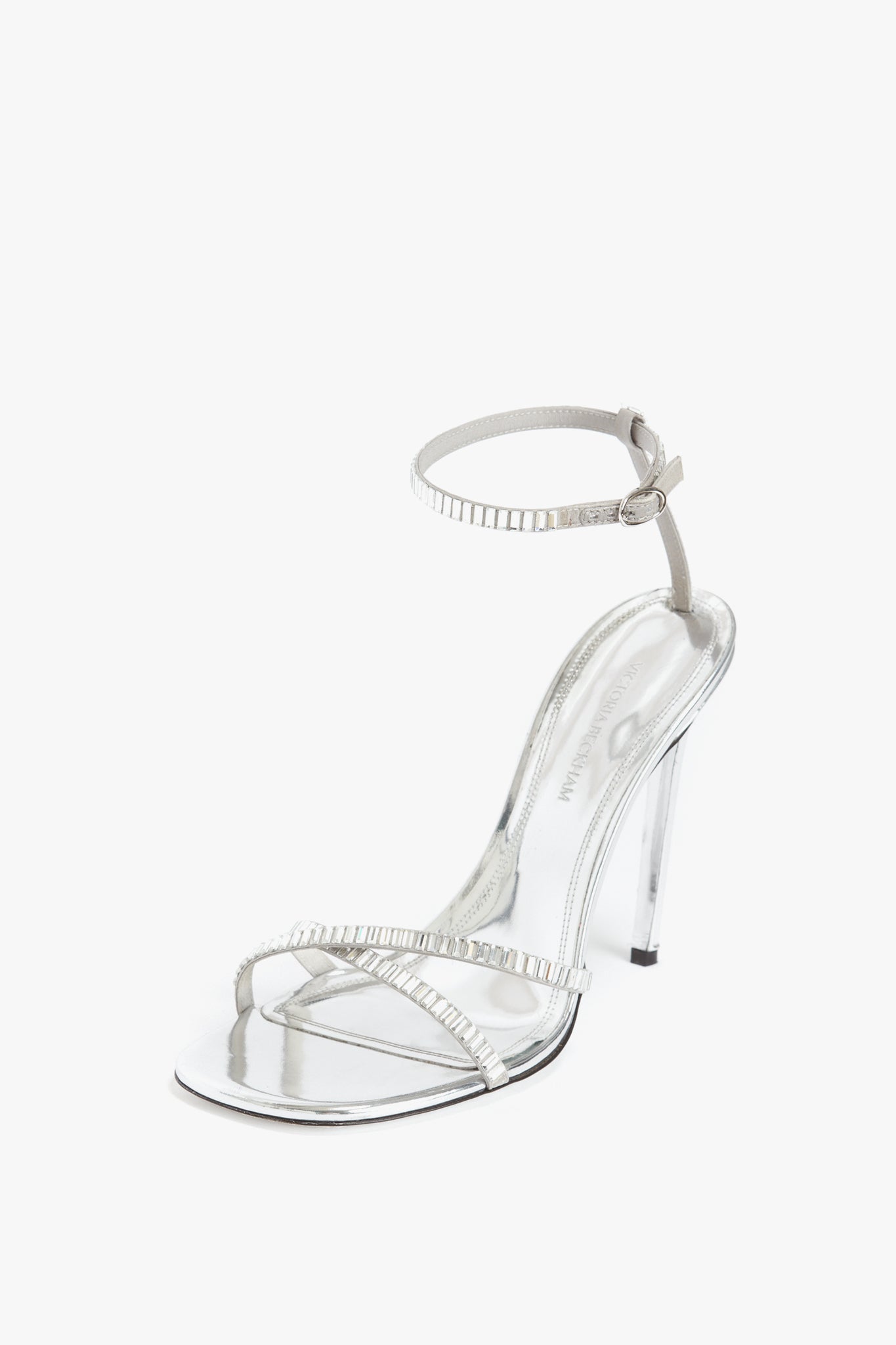 The **Victoria Beckham Rhinestone Sandals In Silver**, featuring an ankle strap and a clear heel, crafted from metallic silver leather.