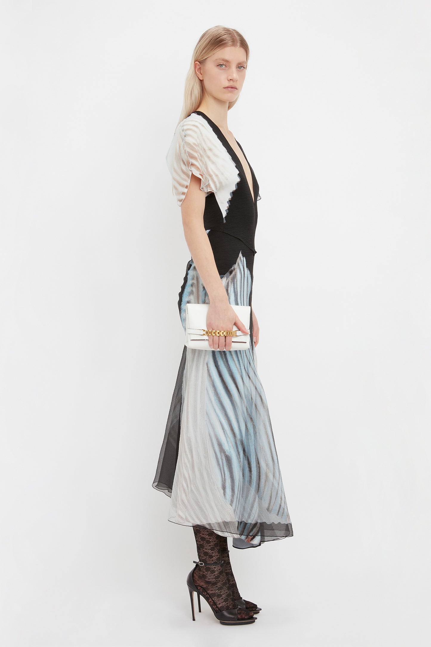 A person stands in profile wearing a black and white patterned dress, lace socks, and black heels, holding a Victoria Beckham Mini Chain Pouch Bag In White Leather. The background is plain white.