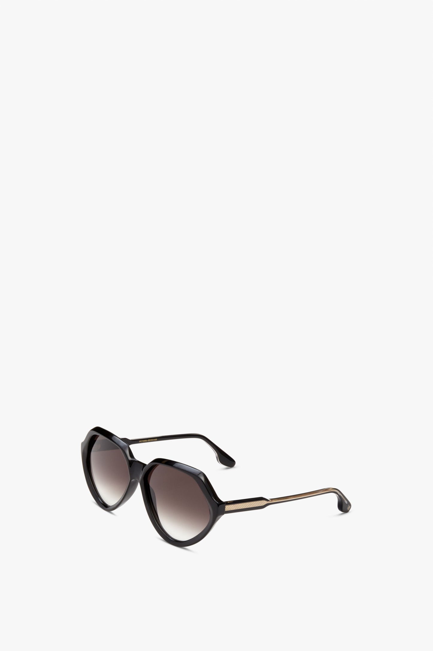 A pair of black, hexagonal-shaped sunglasses with dark tinted lenses and gold accents on the arms, displayed on a plain white background. Faceted Round In Black by Victoria Beckham.