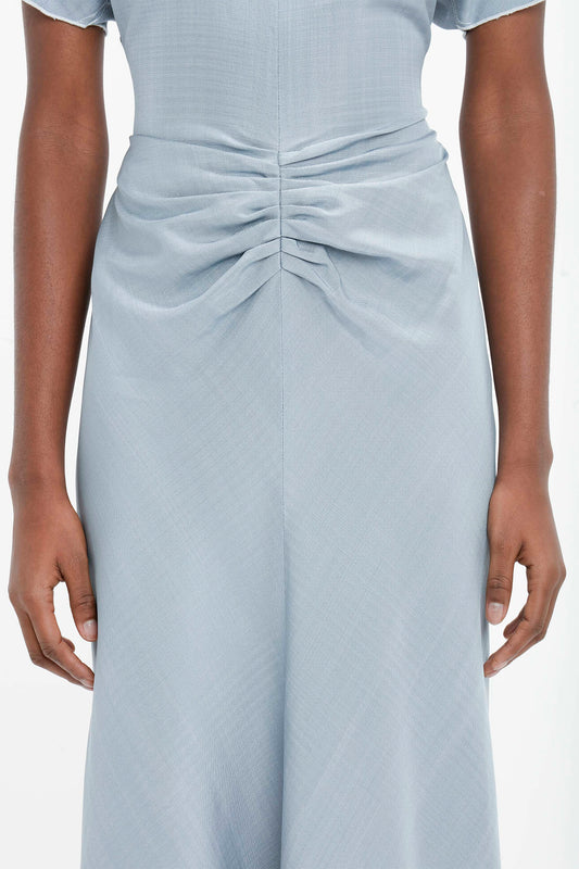 A close-up of a woman's back wearing a light blue dress with a twisted detail at the waist and fit-and-flare silhouette, hands resting gently on her hips - Victoria Beckham's Exclusive Gathered Waist Midi Dress In Pebble.