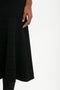 A person wearing a Victoria Beckham Fit And Flare Midi Skirt In Black is partially visible, with their left hand resting on the skirt. The background is plain white.