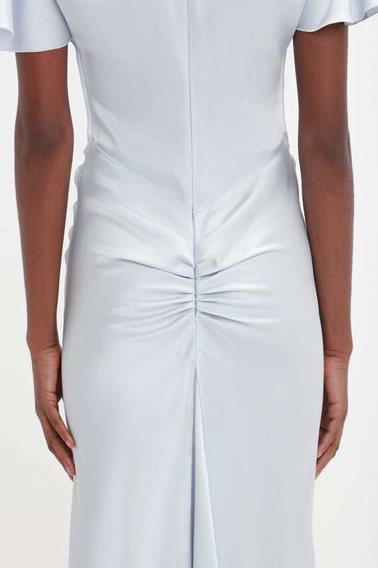 Back view of a person wearing an elegant, light blue Gathered Sleeve Midi Dress In Ice by Victoria Beckham with ruched detailing and a central seam on the lower back. The crepe back satin dress features short sleeves and a smooth, shiny fabric.