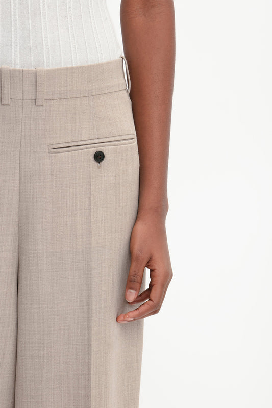 A person is shown from the waist down, donning Victoria Beckham's Reverse Front Trouser In Sesame and a white top, with their right hand resting by their side. The contemporary silhouette highlights their versatile styling.