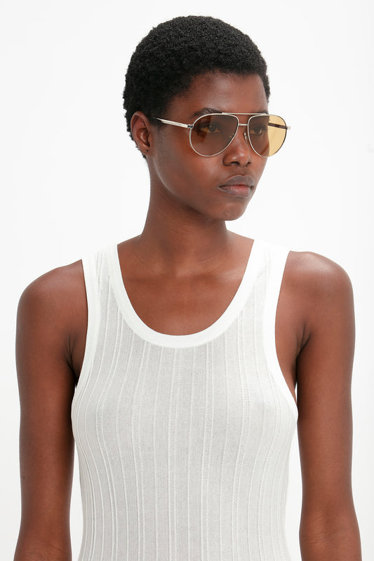 A woman with short hair wearing Victoria Beckham V Metal Pilot Sunglasses in Silver-Brown and a white tank top, standing against a white background.