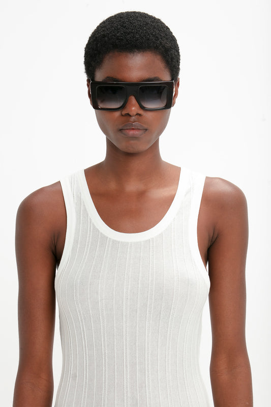 A person with short hair wears Victoria Beckham Oversized Frame Sunglasses In Black and a sleeveless white top with vertical stripes, standing against a plain background, echoing the chic elegance of the SS24 runway.