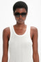 A person wearing Soft Square Frame Sunglasses In Striped Khaki Havana from Victoria Beckham and a sleeveless white top stands against a plain background.