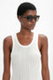 A person wearing a white sleeveless top and large, dark Victoria Beckham Soft Square Frame Sunglasses In Striped Khaki Havana stands against a plain background.