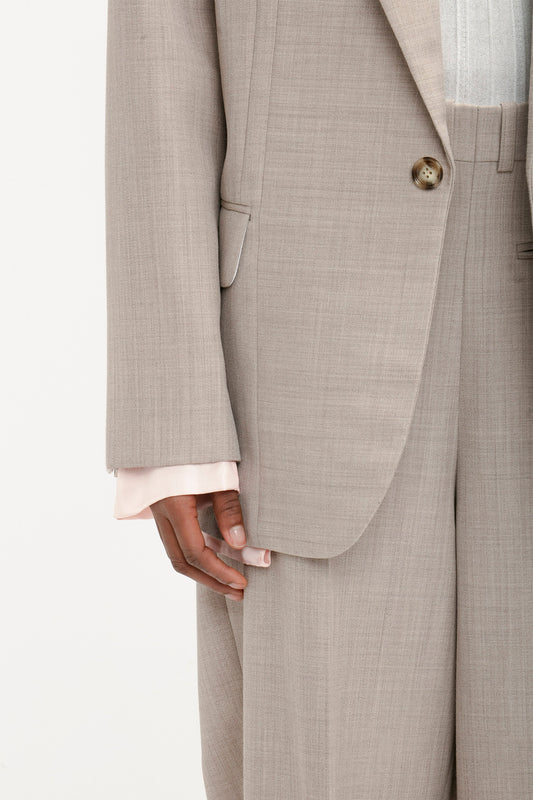 Close-up of a person's hand resting beside their leg, wearing a beige suit with a Darted Sleeve Tailored Jacket In Sesame by Victoria Beckham. A light pink shirt cuff peeks out from under the jacket sleeve, showcasing contemporary detailing.