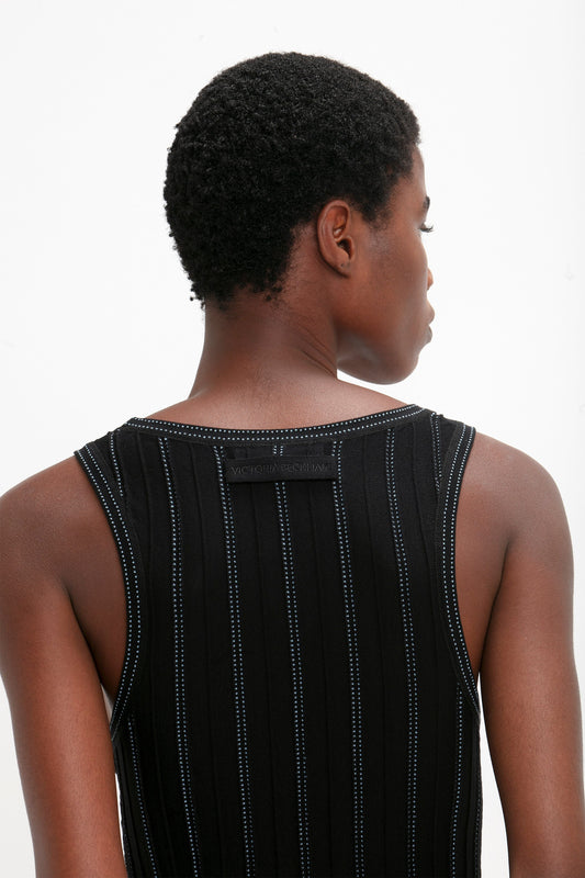 A person with short curly hair is shown from the back, wearing a sleeveless black top with vertical stripes reminiscent of Victoria Beckham's iconic style, featuring a visible tag near the neckline. The top is the Fine Knit Vertical Stripe Tank In Black-Blue by Victoria Beckham.