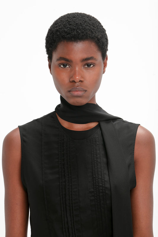 A person with short curly hair is wearing a Sleeveless Tie Neck Top In Black by Victoria Beckham and a luxurious double neck tie scarf around the neck, facing the camera against a plain white background.