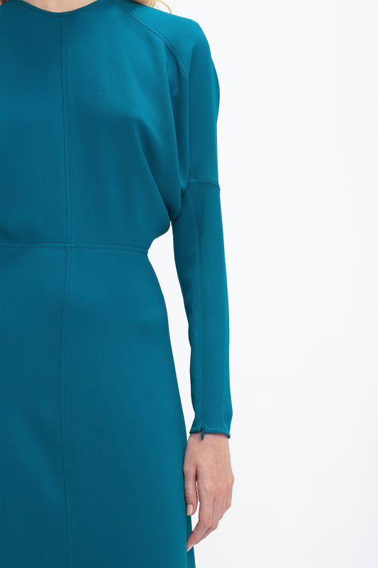 A person wearing a teal Victoria Beckham Long Sleeve Dolman Midi Dress In Petroleum, crafted from fluid cady fabric, is partially visible, with the focus on the upper body and right arm. The background is plain white, enhancing the dress's contemporary sophistication.