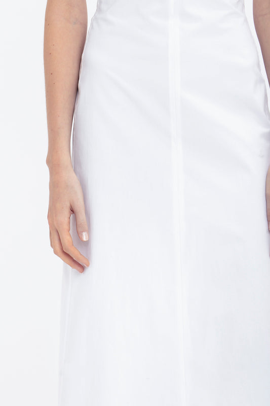 Person wearing a white Cami Fit And Flare Midi In White by Victoria Beckham seen from the shoulders to the mid-thigh. The left arm is visible and relaxed to the side. Made from breathable stretch cotton, this dress features adjustable straps for added comfort. The background is plain white.