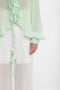 Close-up of a person wearing a light green, Victoria Beckham Romantic Blouse In Jade with ruffled, diaphanous blouson sleeves and white, sheer pants, standing against a plain white background. Only the torso and an arm are visible.