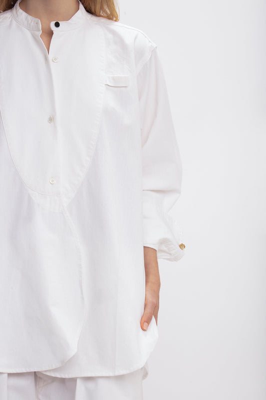 A person is wearing a long-sleeved, relaxed fit, 100% cotton Bib-Front Tuxedo Shirt In Washed White by Victoria Beckham with a mandarin collar and bib-front detailing. Their left hand is in their pocket. The background is plain white.