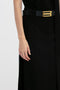 Close-up of a person's left hand resting beside their black outfit with a gold-buckled black belt and a structured silhouette against a white background, featuring the Tailored Floor-Length Skirt In Black by Victoria Beckham.