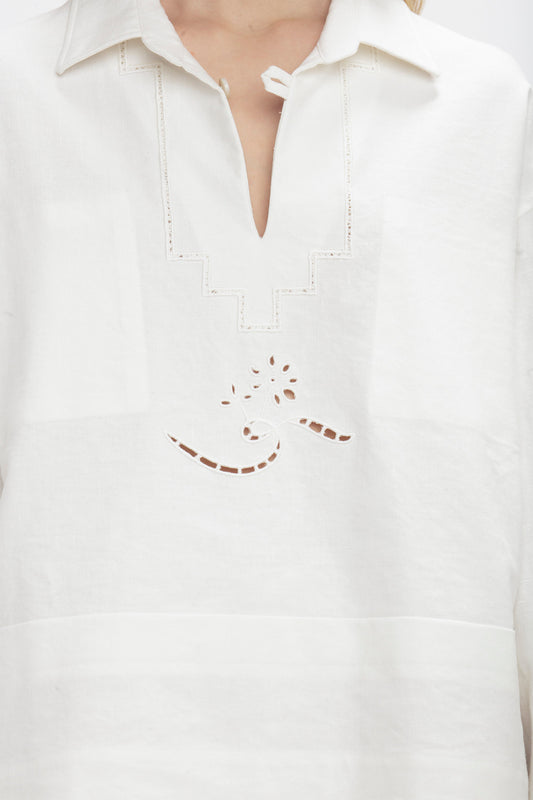 Close-up of a person wearing the Victoria Beckham Oversized Embroidered Tunic In Antique White, featuring a patterned cutout design on the front and a V-neck collar.