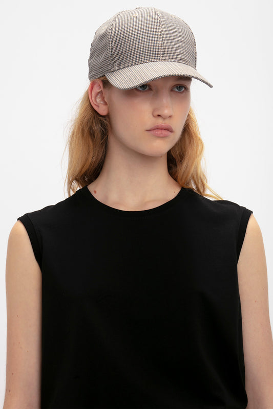 A young woman wearing a black sleeveless top and a gray Victoria Beckham Logo Cap In Dogtooth Check, looking slightly to the side on a plain background.