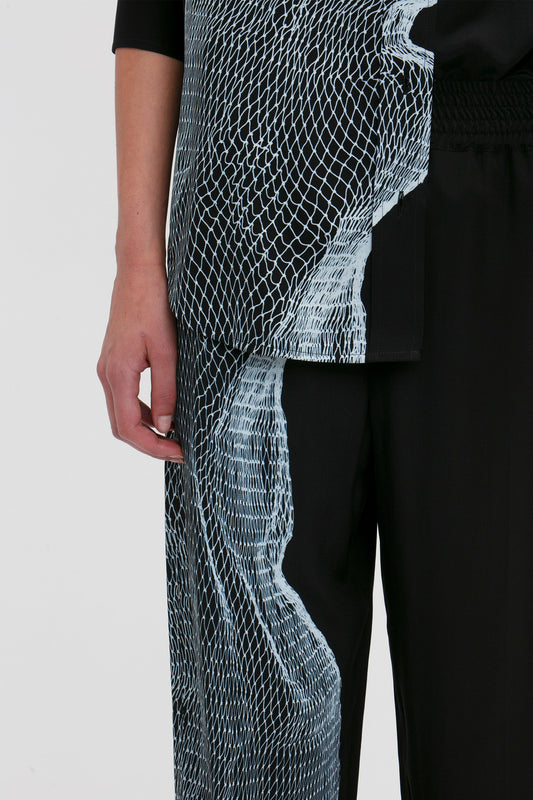 Close-up of a person wearing Victoria Beckham Pyjama Trouser In Black-White Contorted Net with a twisted net print on one side, extending from the top to the pants. Only the left arm and torso are visible against a plain background.