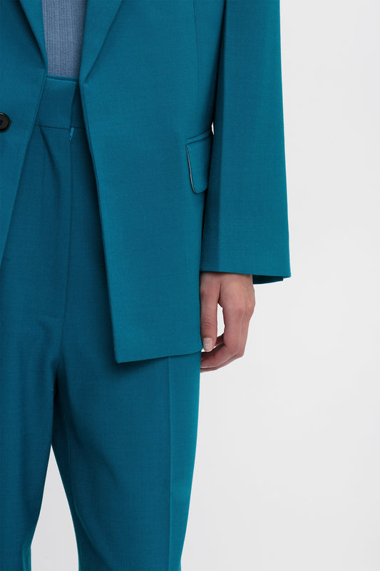 A person wearing a teal, precision-tailored Peak Lapel Jacket In Petroleum with a black button and a light blue shirt is shown from the shoulders down. Their right arm is relaxed at their side, showcasing the elegance reminiscent of Victoria Beckham's design.