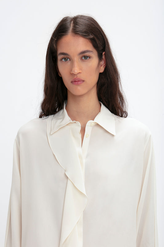 A person with long brown hair wearing a Victoria Beckham Asymmetric Ruffle Blouse In Ivory stands in front of a plain white background.