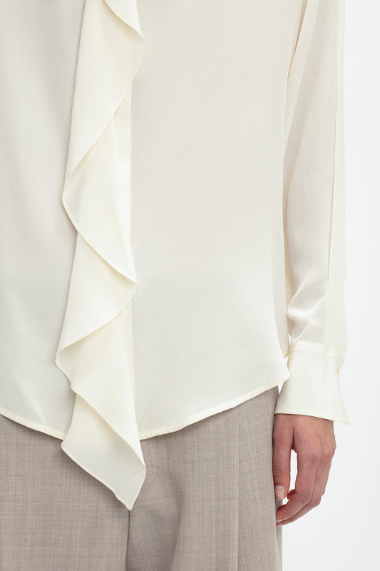 Close-up of a person wearing the Victoria Beckham Asymmetric Ruffle Blouse In Ivory with button-front closure, paired with beige trousers. The person's hand is visible on the side.