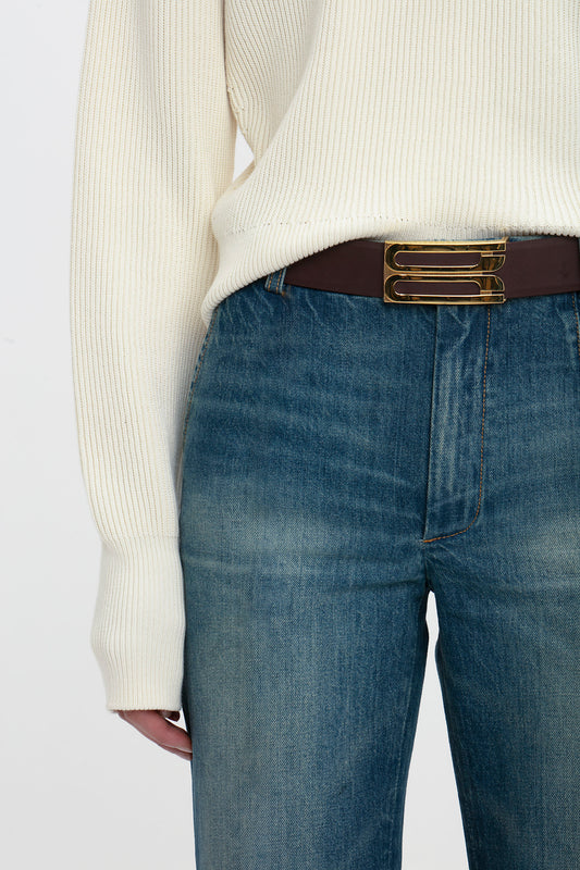 Close-up of a person wearing Victoria Beckham's Cropped Kick Jean In Indigrey Wash and a cream ribbed sweater tucked in with a dark brown belt with a gold buckle.