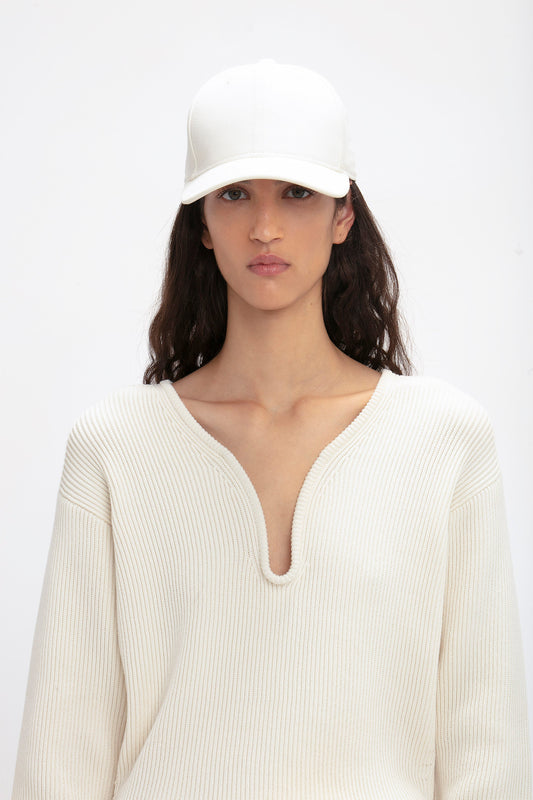 A woman with medium-length dark hair wearing a white Victoria Beckham Logo Cap In Antique White and a ribbed v-neck sweater, looking directly at the camera against a plain background.