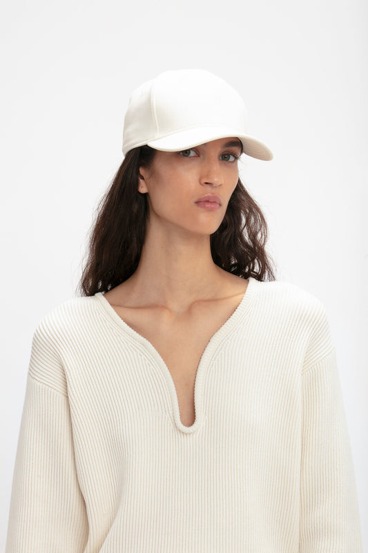 A woman with long brown hair wearing a Victoria Beckham Logo Cap In Antique White and a white ribbed sweater, standing against a plain background, looks directly at the viewer.